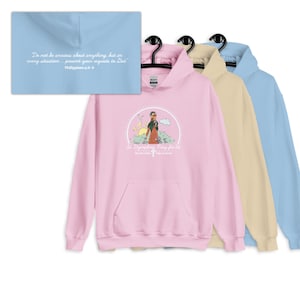 Puede incluir: Tres sudaderas con capucha de color rosa, beige y azul claro con un gráfico de una mujer caminando por un campo con una cita de Filipenses 4:6-8. La cita dice "No se inquieten por nada; más bien, en toda ocasión, con oración y ruego, presenten sus peticiones a Dios, dando gracias."