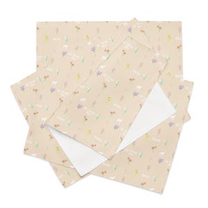 Può includere: Carta da regalo beige con un motivo ripetuto di fiori colorati, agnelli bianchi e il testo "Behold All Things New". Diversi fogli sono sovrapposti, rivelando un interno bianco.