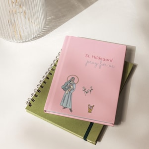 Puede incluir: Un diario rosa con el texto "St. Hildegard pray for us" y una ilustración de una santa. Está apilado sobre un cuaderno verde. En el fondo se ve un jarrón de cristal transparente.