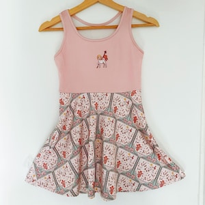 Może przedstawiać: Różowa sukienka bez rękawów z haftowanym wzorem w kolorze białym i czerwonym na gorsecie. Spódnica ma wzór z motywem kwiatowym.