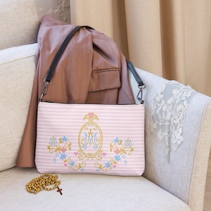 Può includere: Borsa a tracolla a righe rosa e bianche con cinturino nero. La borsa presenta un emblema oro e blu con la lettera "M" e dettagli floreali. Sono visibili anche un rosario marrone e un blazer rosa.