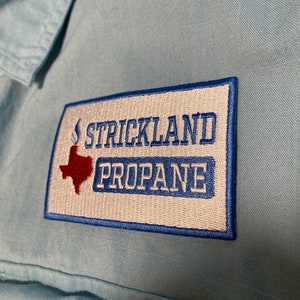 May include: A light blue button-down shirt featuring a rectangular embroidered patch on the chest. The patch has a white background with a blue border, the text STRICKLAND PROPANE in blue, and a red silhouette of the state of Texas with a flame.
