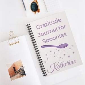 Pode incluir: Um caderno espiralado com o texto "Gratitude Journal for Spoonies" em roxo. Um desenho de colher roxa e o nome "Katherine" também são apresentados. O caderno está aberto, revelando páginas em branco.