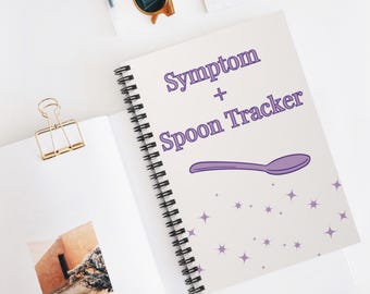 May include: A spiral-bound notebook with the text "Symptom + Spoon Tracker" in purple. A purple spoon graphic is below the text, with purple star accents. The notebook is open, with a gold binder clip and a small photo visible.