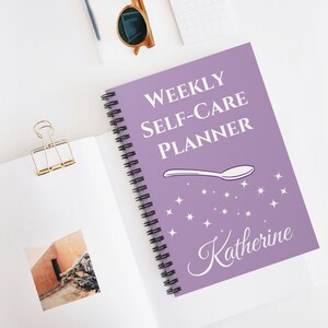 Peut inclure: Un agenda hebdomadaire de soins personnels à spirales, avec une couverture lavande. La couverture affiche le texte "WEEKLY SELF-CARE PLANNER", une illustration de cuillère et le nom "Katherine". L'agenda est ouvert sur une page blanche.