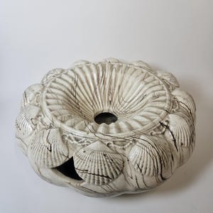 May include: A white ceramic bowl with a seashell design. The bowl is shaped like a large seashell and has a hole in the center. The bowl is decorated with a marbled pattern and has a raised seashell design around the rim.
