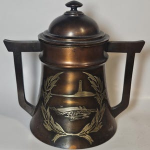 May include: A copper and silver teapot with a lid and two handles. The teapot has a design of a lighthouse and a beach scene on the front.