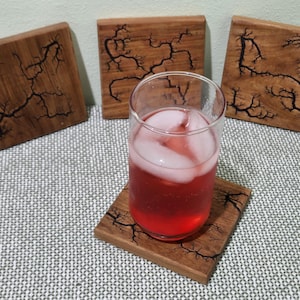 May include: Four square wooden coasters with a dark wood grain finish and a black fractal design. One coaster holds a glass of red liquid with ice. The coasters are on a patterned surface.