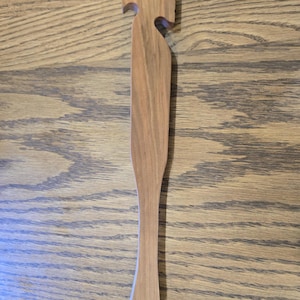 以下が含まれることがあります： ユニークなデザインの木製キッチン用品。明るい茶色のヘラは、湾曲したハンドルと切り欠きのある装飾的な上部が特徴です。ハンドルの端には小さな穴があります。この器具は木製です。