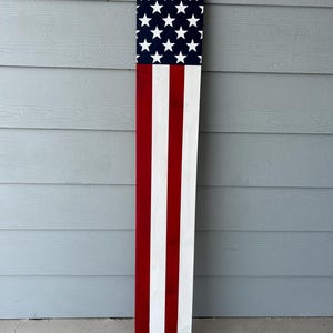 May include: A wooden American flag decoration. The top section features a blue background with white stars, while the lower section displays red and white stripes. The flag is rectangular and stands upright.
