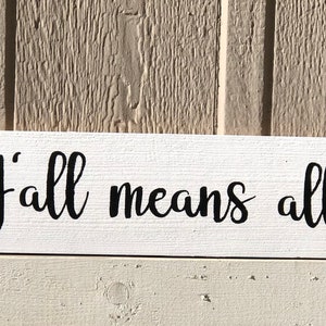 May include: White wooden sign with the text "Y'all means all" in black cursive lettering. A rainbow heart is on the left side of the sign and a light blue and pink transgender pride heart is on the right side.