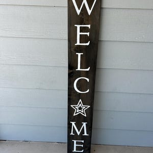 May include: A dark brown wooden welcome sign with white lettering and a star design. The sign is rectangular with a unique top shape and is designed for outdoor use. The word "WELCOME" is vertically displayed.