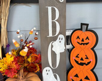 May include: A wooden sign with the words "hey boo" painted in white with ghost silhouettes. The sign is leaning against a stack of three orange pumpkins with black jack-o-lantern faces.