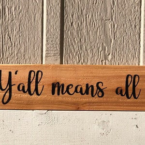 May include: Wooden sign with the text "Y'all means all" in black cursive lettering. A rainbow heart is on the left and a transgender pride heart is on the right.