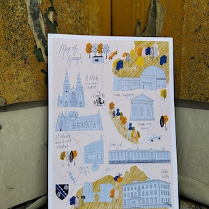 Puede incluir: Un mapa dibujado a mano de Armagh, Irlanda del Norte, con acentos azules y amarillos. El mapa presenta puntos de referencia como la Catedral de San Patricio, el Planetario de Armagh y el Museo del Condado de Armagh. El texto "Map of Armagh" está escrito en cursiva azul en la parte superior del mapa.