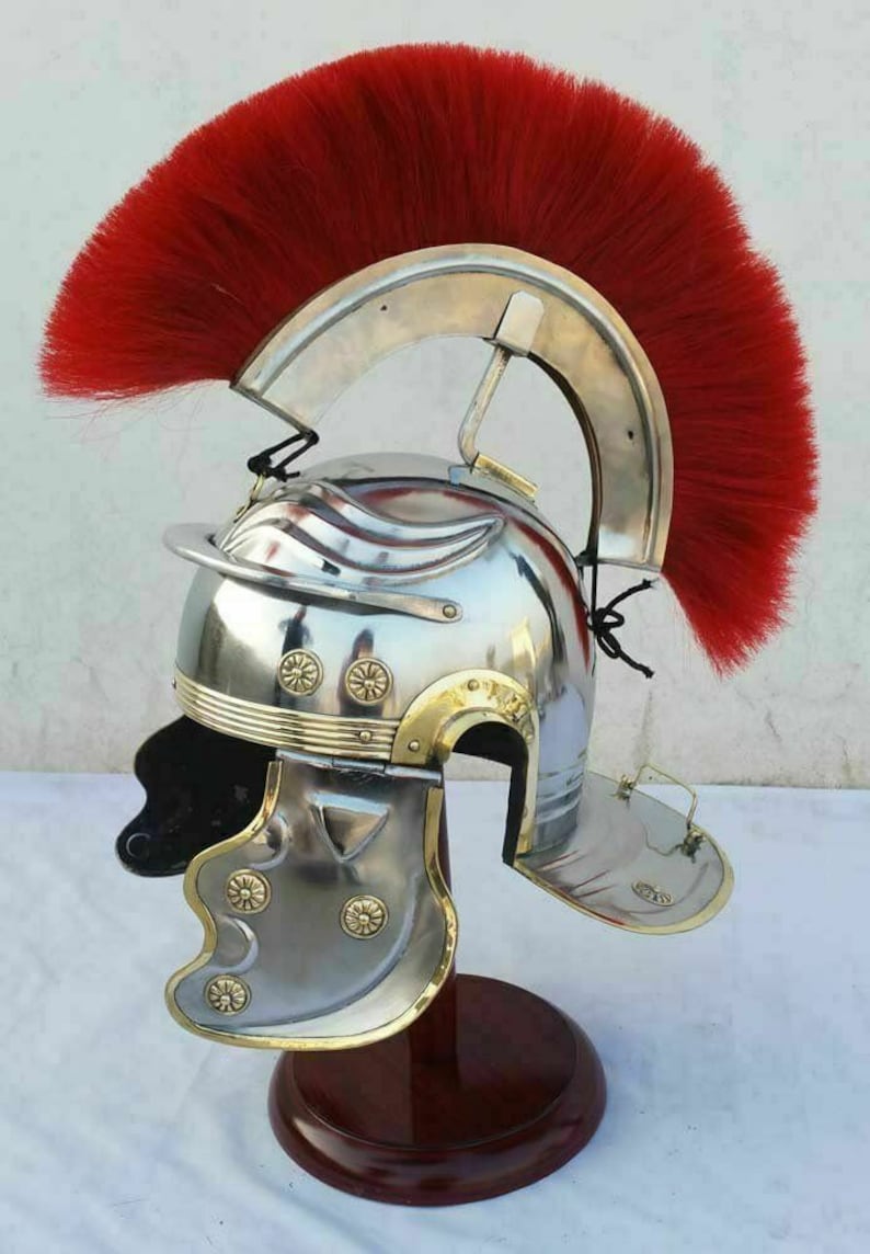 May include: A silver Roman helmet with a red horsehair crest, brass accents, and a cheek guard. The helmet is displayed on a wooden stand.