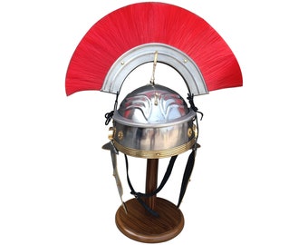 May include: A silver Roman helmet with a red plume on a wooden stand. The helmet has a brass trim and a decorative design on the top.