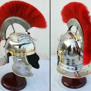 May include: A silver Roman helmet with a red plume, featuring a brass trim and decorative details. The helmet is displayed on a wooden stand.
