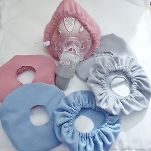 May include: Four fabric face mask covers in blue and pink. Two of the covers are round with a hole in the center. The other two covers are gathered around the hole in the center. A clear plastic face mask with a pink fabric cover is also in the image.