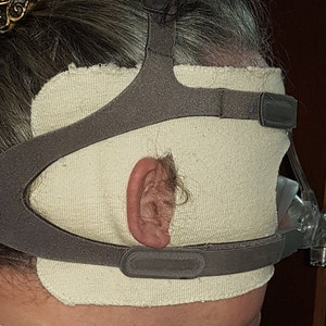 Puede incluir: Primer plano de una máscara para dormir de tela beige con una correa gris y una hebilla negra. La máscara se lleva sobre la oreja de una persona.