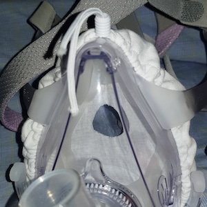 May include: A clear plastic CPAP mask with a white fabric liner and a gray head strap. The mask has a clear plastic tube attached to it.
