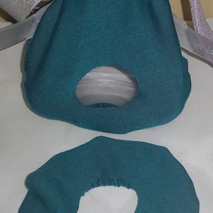 May include: Two teal blue fabric face mask covers. The top cover has a hole in the center. The bottom cover is a flat circle with a hole in the center.