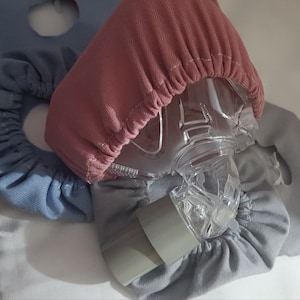 May include: A set of three fabric covers for a CPAP mask. The covers are in shades of blue and pink. The covers are made of a soft, stretchy fabric and are designed to fit snugly over the mask.