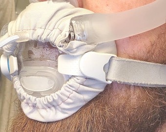 May include: A CPAP mask with a clear plastic face covering, white straps, and a soft, light gray fabric cover. The mask is designed to fit over the nose and mouth, with a flexible tube attached for airflow.