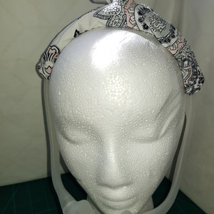 May include: A white headband with a pink and black paisley pattern. The headband is worn on a white mannequin head. A clear plastic nasal cannula is attached to the headband.