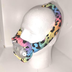 Puede incluir: Una mascarilla CPAP con una funda de tela con estampado de leopardo arcoíris. La mascarilla está sobre una cabeza de maniquí blanca. La tela tiene una mezcla de colores, incluyendo amarillo, naranja, rosa, azul y manchas negras. La mascarilla tiene un componente de plástico transparente.
