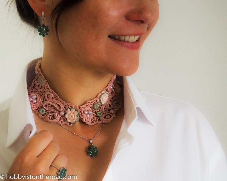K&ouml;nnte beinhalten: Ein rosa H&auml;kelkragen mit gr&uuml;nen und wei&szlig;en Akzenten. Der Kragen wird &uuml;ber einem wei&szlig;en Hemd getragen und mit einer silbernen Kettenkette mit einem gr&uuml;nen Anh&auml;nger und Ohrringen kombiniert.