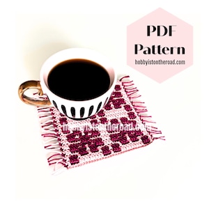 May include: A white teacup with a gold handle filled with dark coffee sits on a handmade coaster. The coaster is square, with a burgundy and white geometric pattern and fringe. Text on the image reads "PDF Pattern" and "hobbyisontheroad.com".