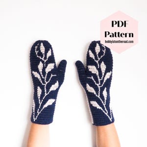 May include: A pair of navy blue crocheted mittens with a white leaf design. The mittens are shown on a white background. PDF Pattern hobbyistontheroad.com