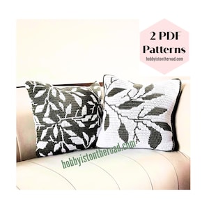 May include: Two square crochet pillows with a leaf design. One pillow is white with dark gray leaves, and the other is dark gray with white leaves. The pillows are on a white couch. Text on the image reads "2 PDF Patterns" and "hobbyisontheroad.com".