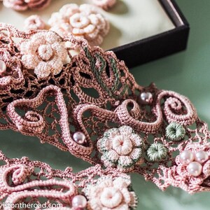 K&ouml;nnte beinhalten: Eine zarte geh&auml;kelte Spitzenborte mit rosa und gr&uuml;nen Blumenmotiven und Perlenakzenten. Die Spitze ist filigran und detailliert, mit einem zarten Blumenmuster.