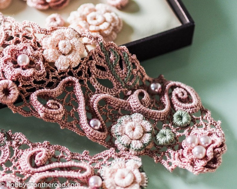 K&ouml;nnte beinhalten: Eine zarte geh&auml;kelte Spitzenborte mit rosa und gr&uuml;nen Blumenmotiven und Perlenakzenten. Die Spitze ist filigran und detailliert, mit einem zarten Blumenmuster.