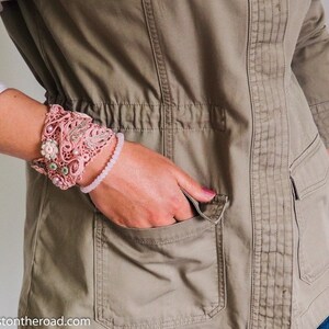 K&ouml;nnte beinhalten: Ein rosa geh&auml;keltes Armband mit Blumenschmuck und ein rosa Perlenarmband am Handgelenk einer Person.