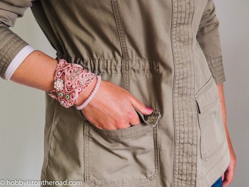 K&ouml;nnte beinhalten: Ein rosa geh&auml;keltes Armband mit Blumenschmuck und ein rosa Perlenarmband am Handgelenk einer Person.
