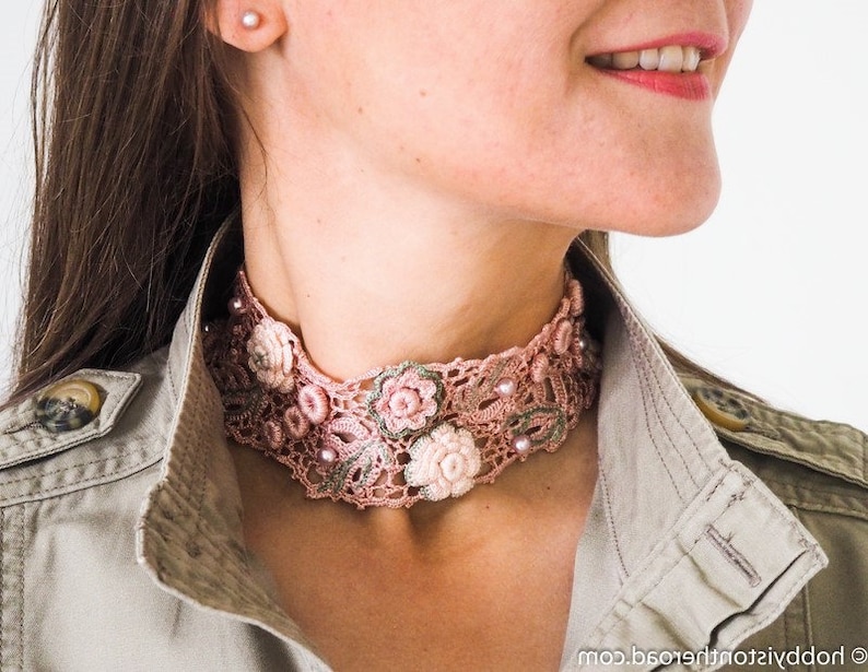 K&ouml;nnte beinhalten: Eine rosafarbene geh&auml;kelte Choker-Halskette mit floralen Verzierungen und Perlenakzenten.