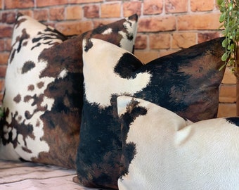 May include: Close-up of three decorative throw pillows with a cowhide pattern. The pillows feature a mix of white, brown, and black patches, creating a rustic and textured appearance. The pillows are arranged in a layered composition, suggesting a cozy home decor theme.