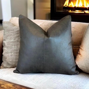Peut inclure: Un coussin de canap&eacute; en similicuir gris fonc&eacute; avec un l&eacute;ger brillant. Le coussin est sur un canap&eacute; gris clair avec d'autres coussins.