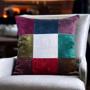 May include: A patchwork cushion featuring a range of colours including burgundy, teal, white, red, and brown. The cushion is made of velvet and has a square shape.