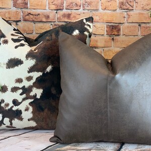 May include: Two decorative throw pillows. One pillow features a cowhide pattern in shades of brown and white. The other pillow is a solid, textured gray-brown. Both pillows are square and have a soft, inviting appearance.