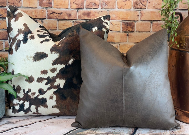 May include: Two decorative throw pillows. One pillow features a cowhide pattern in shades of brown and white. The other pillow is a solid, textured gray-brown. Both pillows are square and have a soft, inviting appearance.
