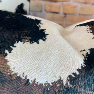 May include: Close-up of a textured pillow with a cowhide pattern. The design features patches of black, brown, and cream colors. The fabric has a woven appearance, and the pillow is slightly curved.