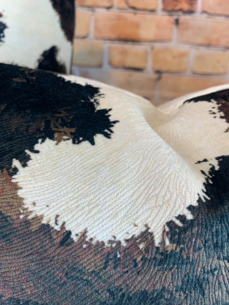 May include: Close-up of a textured pillow with a cowhide pattern. The design features patches of black, brown, and cream colors. The fabric has a woven appearance, and the pillow is slightly curved.