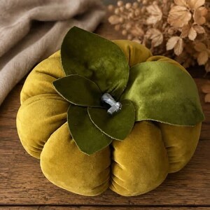 May include: A plush, mustard-yellow velvet pumpkin decoration featuring deep, rounded segments. The top is adorned with several overlapping, dark olive-green velvet leaves and a small, central silver-coloured stem accent.