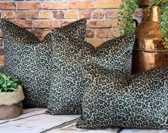 May include: Four decorative throw pillows with a leopard print pattern in shades of tan and black. The pillows are arranged in a group, showcasing their varying sizes. The background includes a brick wall and a copper container.