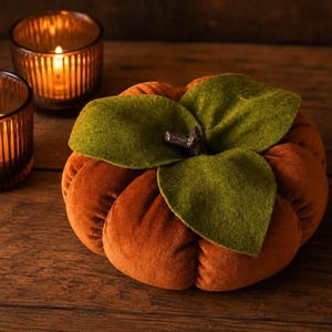 May include: A plush, round, orange velvet pumpkin decoration featuring three dark green felt leaves attached to a central dark brown stem, positioned next to two small, ribbed glass candle holders containing lit tea light candles.