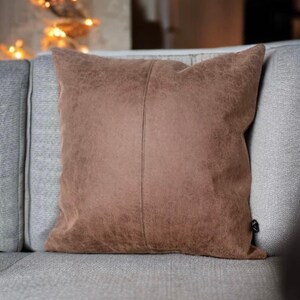 Peut inclure: Un coussin en daim brun avec une couture discr&egrave;te au centre. Le coussin est pos&eacute; sur un canap&eacute; gris clair.